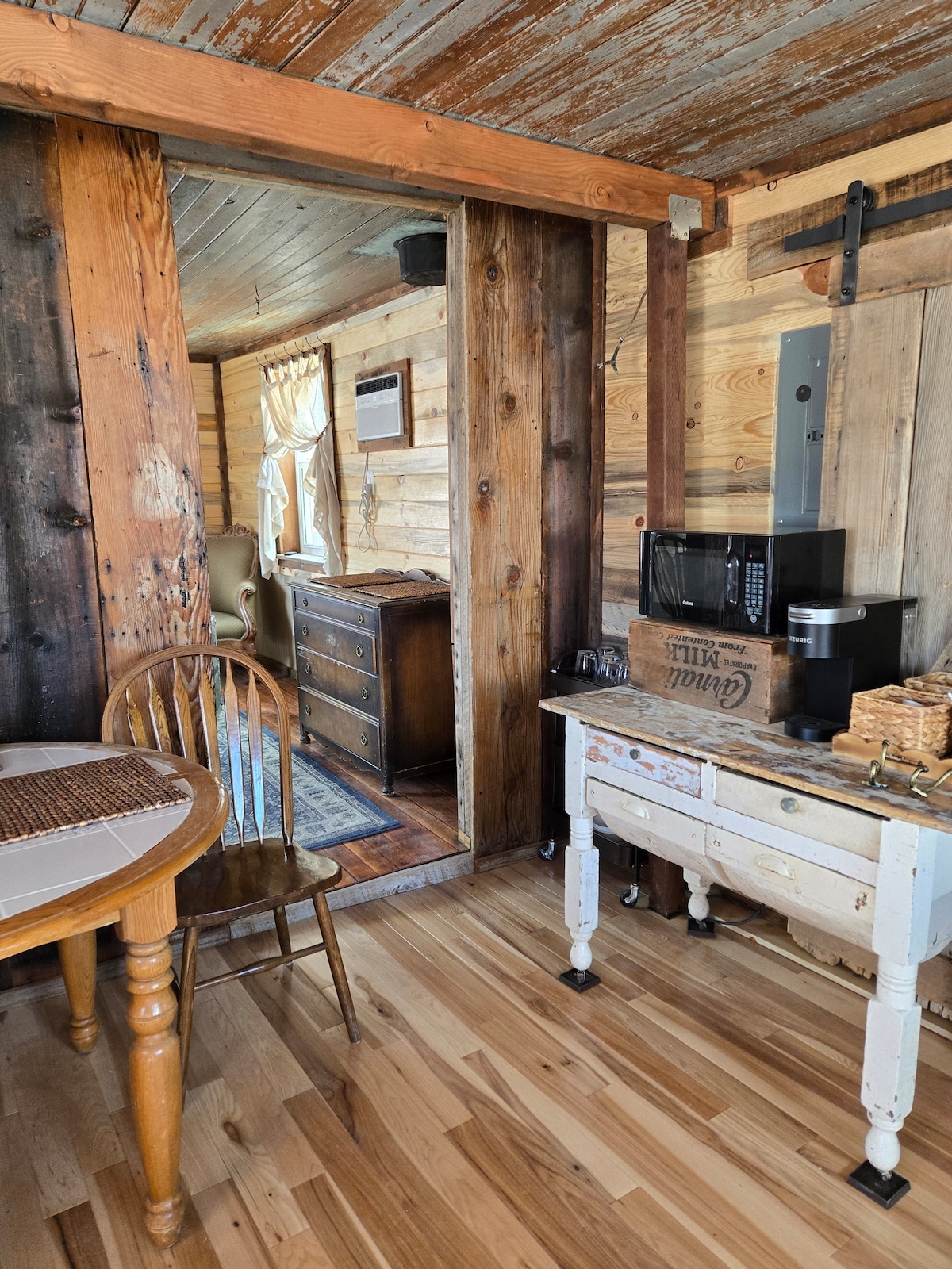 The cabin's interior combines rustic wood accents with a warm, inviting atmosphere. A small dining table with wooden chairs is visible, alongside a vintage-style console table. Nearby, a microwave sits atop the console, and inviting light filters through a window leading to a cozy sitting area.