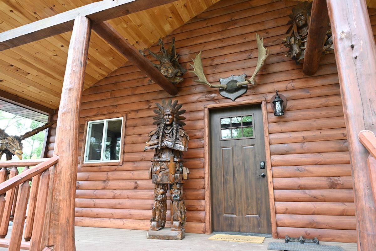 The entrance of a log cabin is highlighted by a wooden door, surrounded by intricate carvings and sculptures. A large decorative figure stands to one side, accentuating the rustic charm. Natural wood beams and a welcoming porch add to the cabin's cozy appeal.