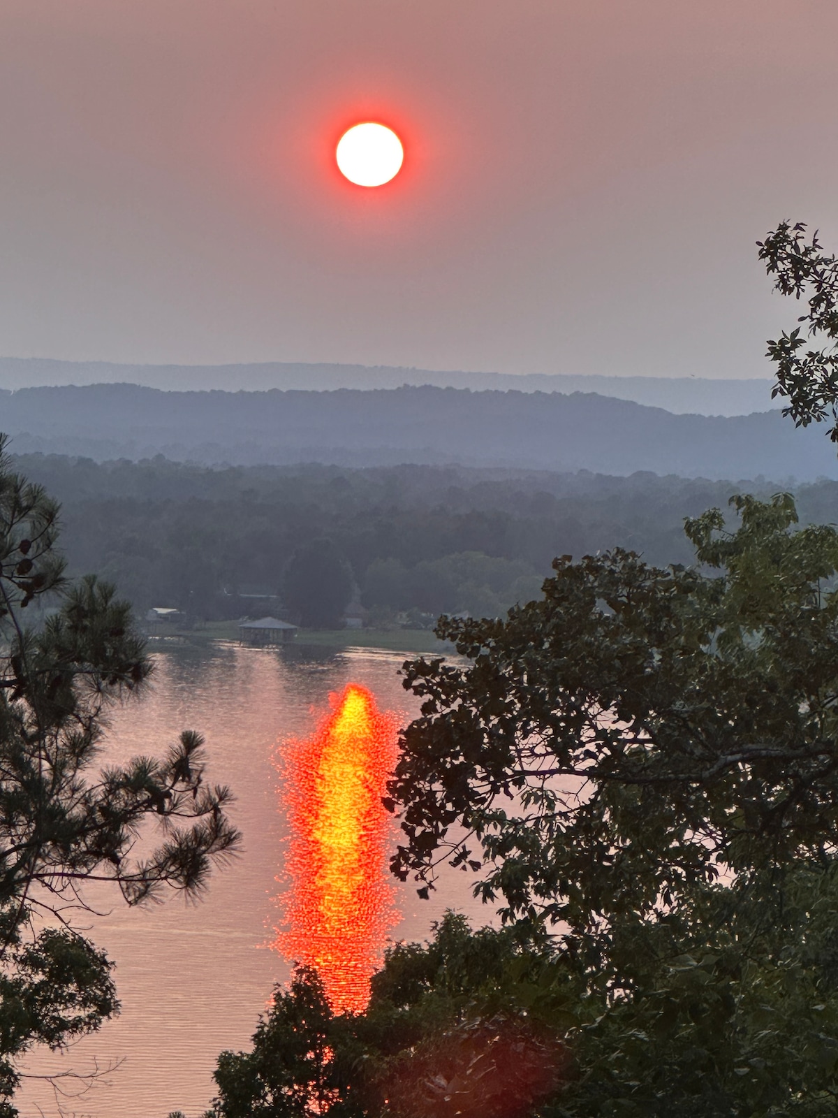 A vibrant sunset is captured over a serene river, with the sun reflecting in bright hues on the water's surface. Soft hills and greenery frame the scene, creating a peaceful backdrop for this natural display.