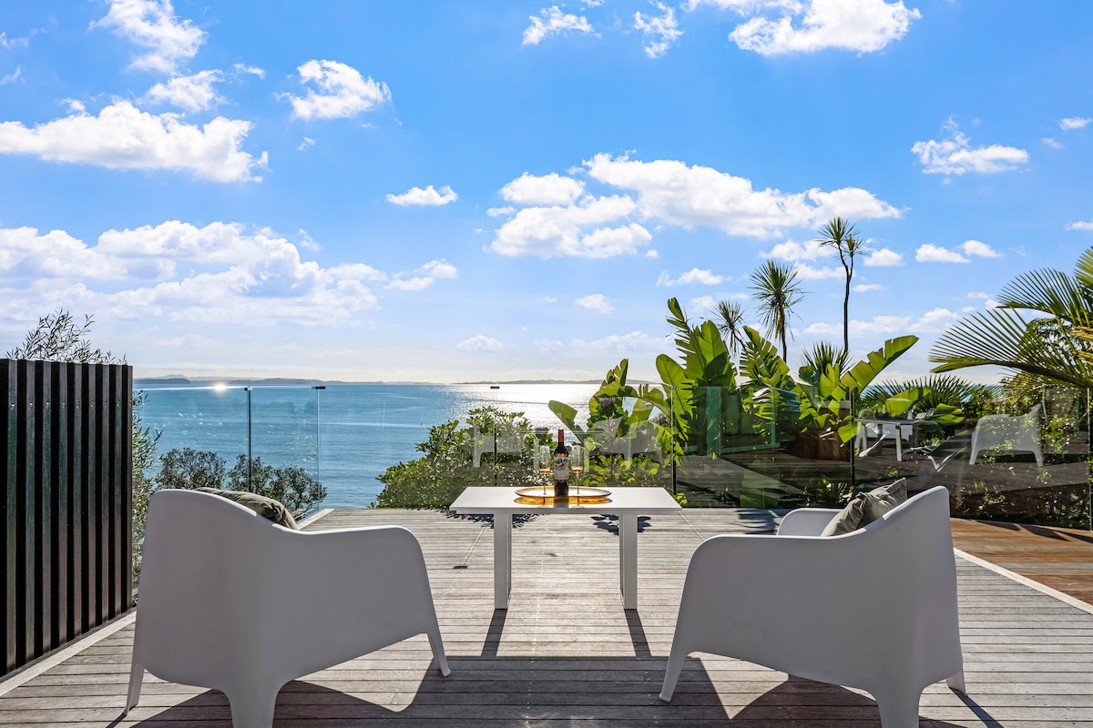 A spacious outdoor deck features modern white seating and a small table, inviting relaxation while overlooking the expansive sea. Lush greenery surrounds the area, and the bright sky complements the shimmering water in the distance.