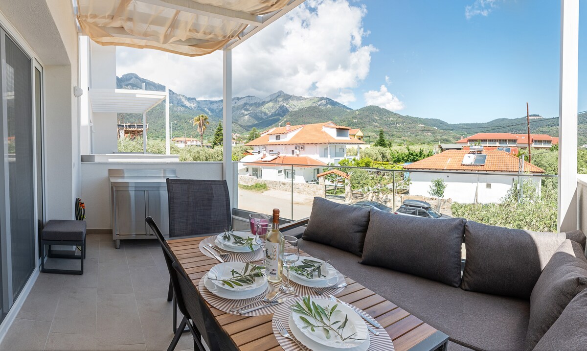 A spacious balcony offers a wooden dining table set with decorative plates and glasses. Comfortable seating with gray cushions is arranged around the table, providing a relaxed atmosphere. The backdrop features distant mountains and a view of the surrounding village, with clear blue skies above.