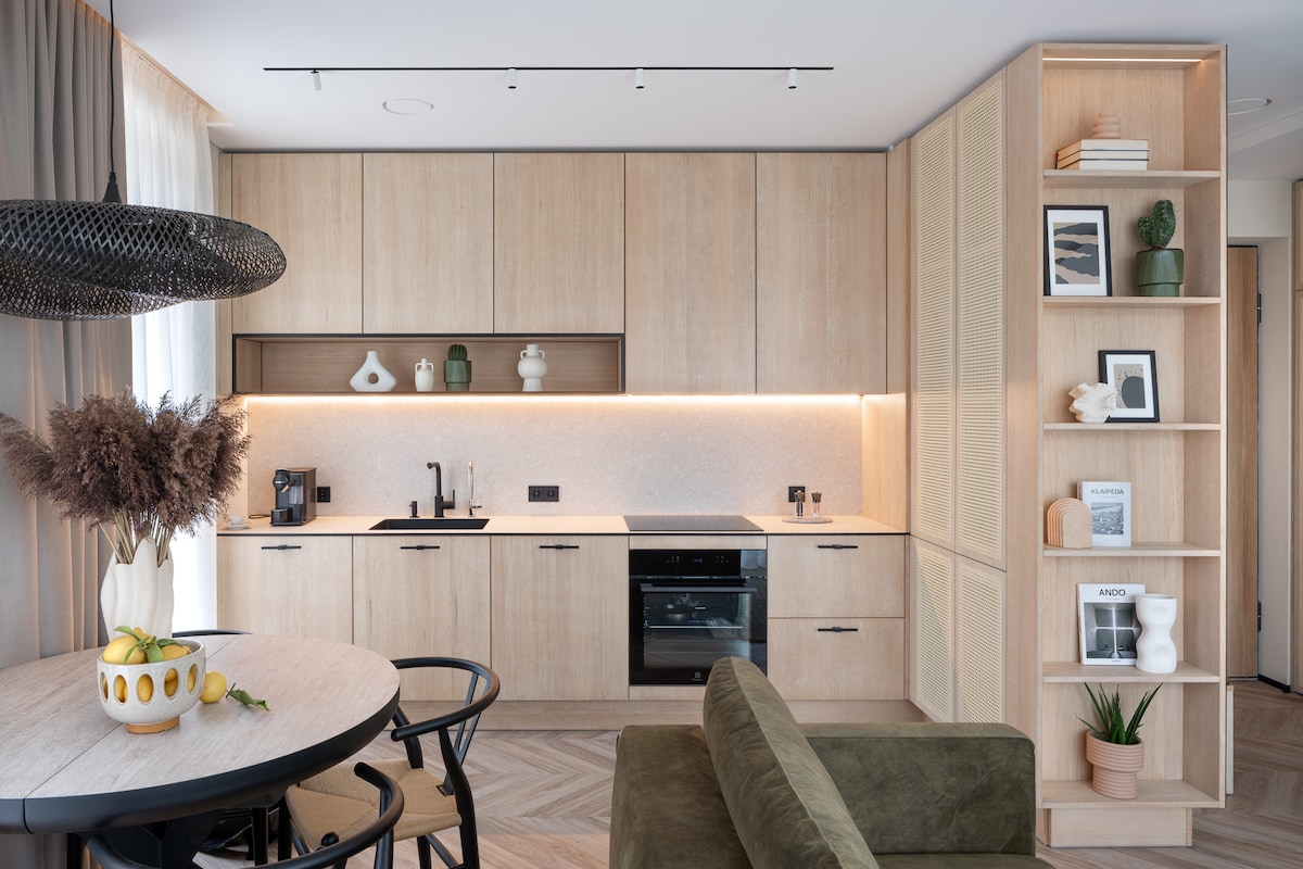 A modern kitchen showcases wood cabinetry with subtle lighting, complemented by a sleek built-in oven and countertop appliances. A round dining table with decorative items and a fruit bowl is positioned nearby. Soft furnishings can be seen in the living area, enhancing the inviting space.