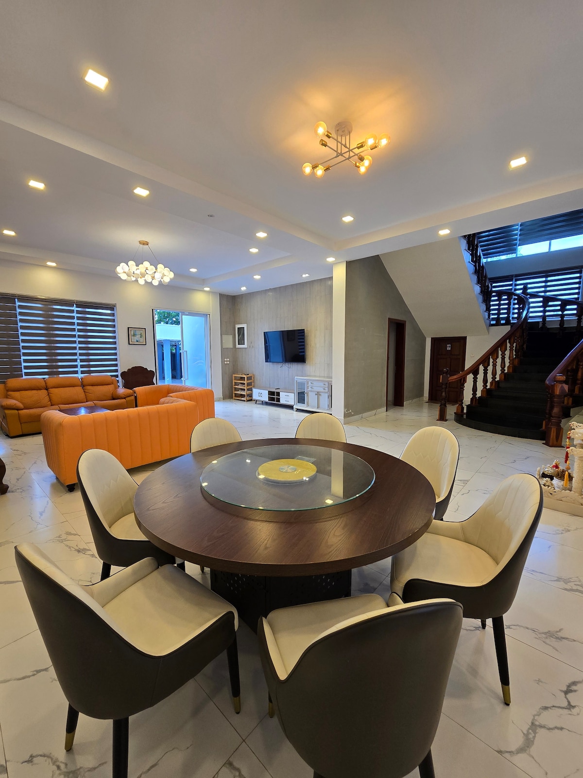 A spacious living area features an inviting orange sofa and a round dining table surrounded by six chairs. Natural light fills the room through large windows, with a staircase leading to the upper level. Decorative elements include a wall-mounted television and soft ceiling lighting.