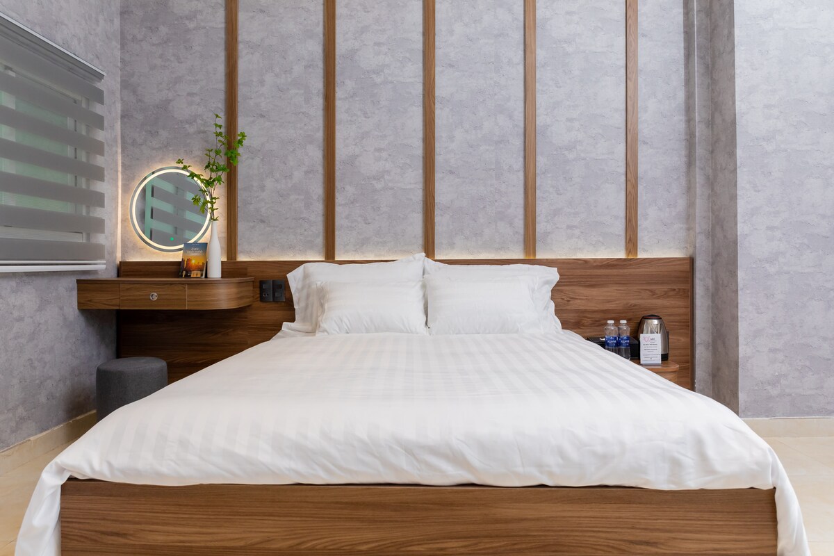 A spacious bed is positioned against a light grey wall, featuring a contemporary design with wooden accents. A small bedside table is visible, accompanied by a minimalistic mirror and a potted plant. Bottled water is placed on the side, creating a functional and clean aesthetic.