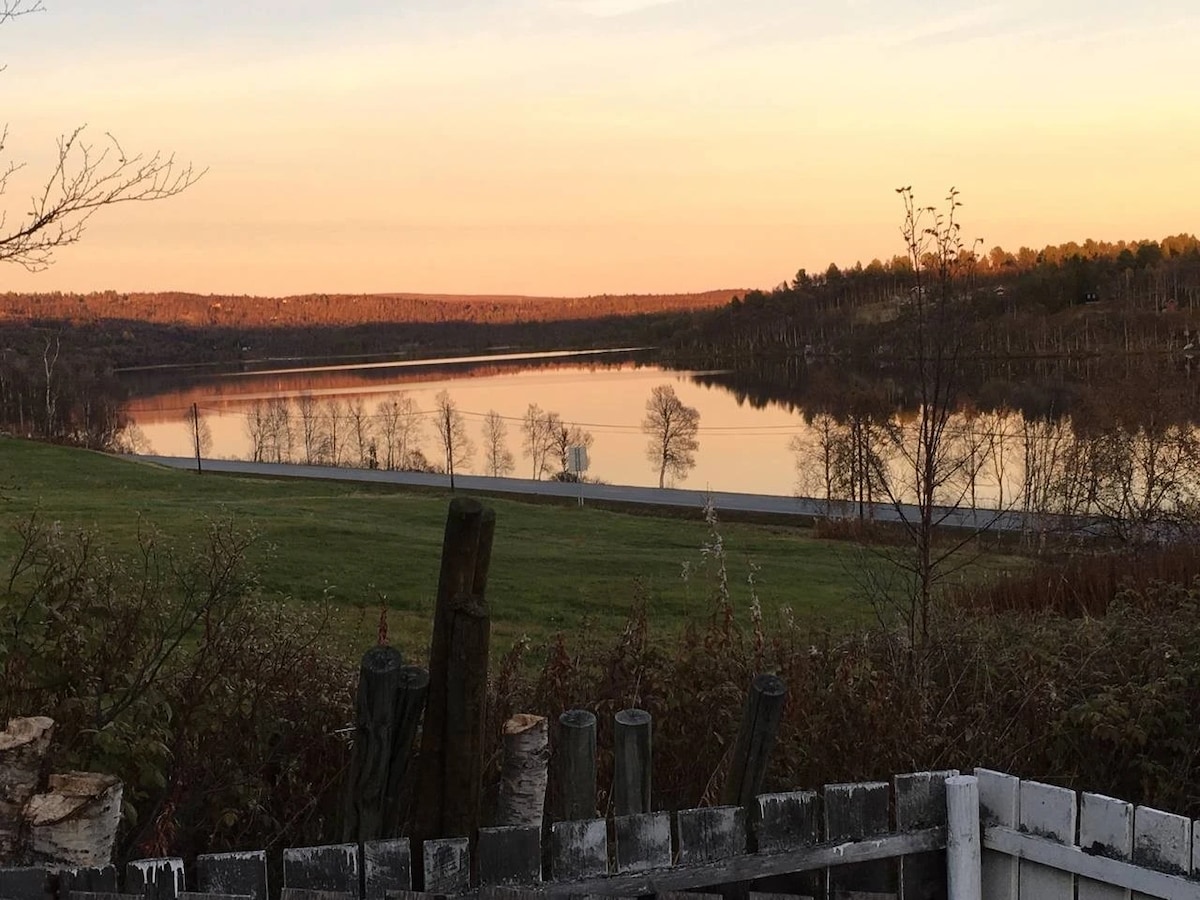 A serene view of Lake Hittersjoen is captured at sunset, with soft hues reflecting on the water's surface. Surrounding grassy areas and distant trees create a peaceful natural setting, enhancing the visual experience.