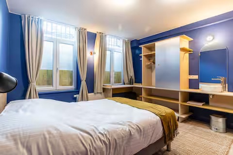 A cozy private bedroom is presented, featuring a spacious bed with light linens and a mustard-colored throw. Natural light enters through two windows, complemented by sheer curtains. A minimalist wardrobe and a small sink with a mirror are positioned against a blue accent wall.