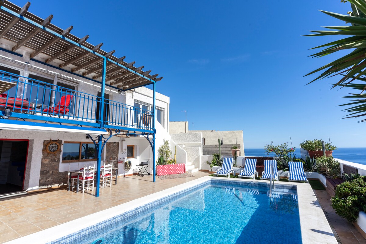 A private pool area is highlighted under clear blue skies, surrounded by sun loungers. The two-story villa features a spacious terrace with a blue railing, red chairs, and lush greenery. The outdoor dining area is visible to the left, enhancing the inviting atmosphere.