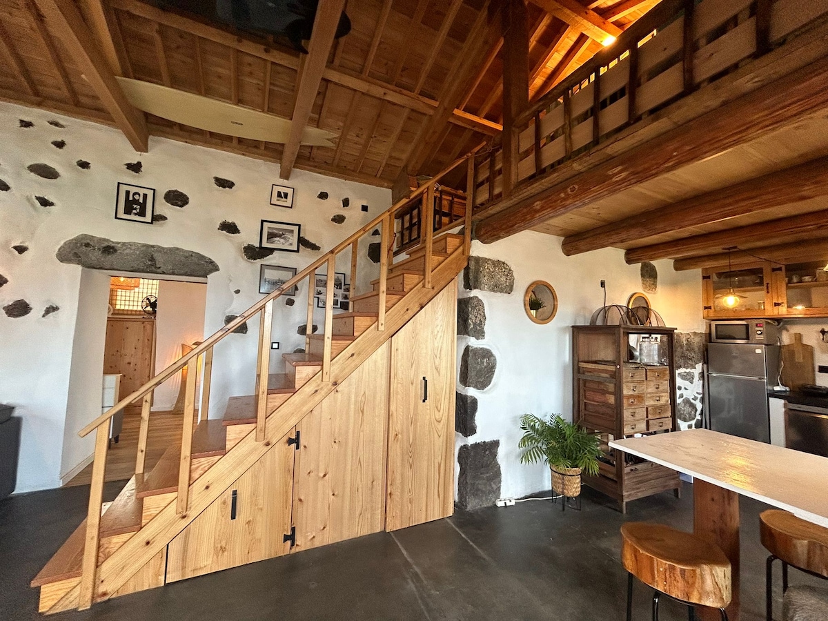 The interior space features an open layout with an inviting wooden staircase leading to a mezzanine level. Stone walls are complemented by wooden beams and accents. A cozy dining area is visible, along with a well-equipped kitchen and decorative elements that enhance the natural ambiance.