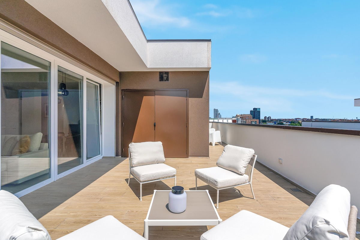 An expansive private terrace is furnished with white outdoor chairs and a small table. The flooring is wooden, contributing to a modern aesthetic. Large sliding glass doors provide access to the interior and allow natural light to fill the space. The surrounding views showcase a clear sky.