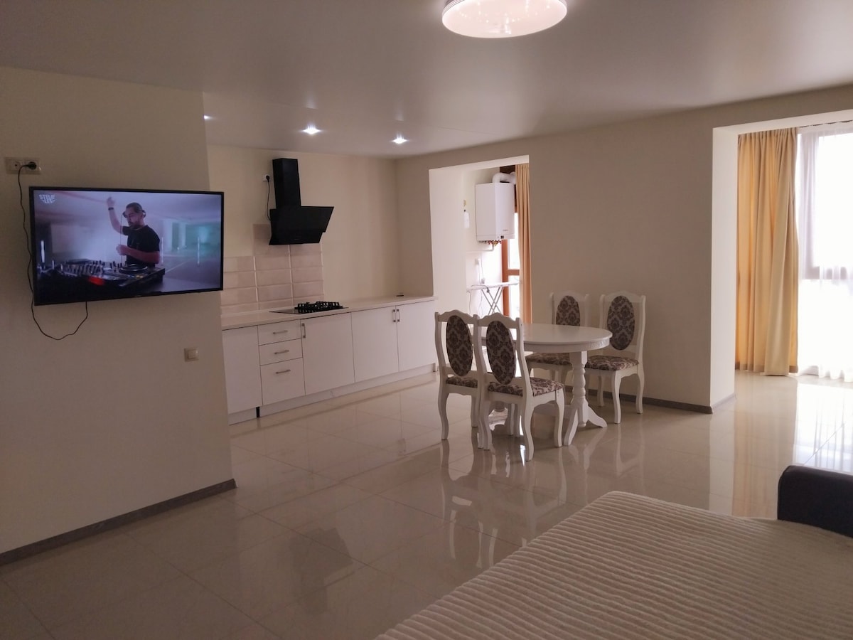 A modern living area features a wall-mounted television displaying an entertainment scene. A round dining table with four chairs is positioned near the kitchen area, which includes sleek cabinets and a stovetop. Natural light enters from a window, highlighting the polished flooring.