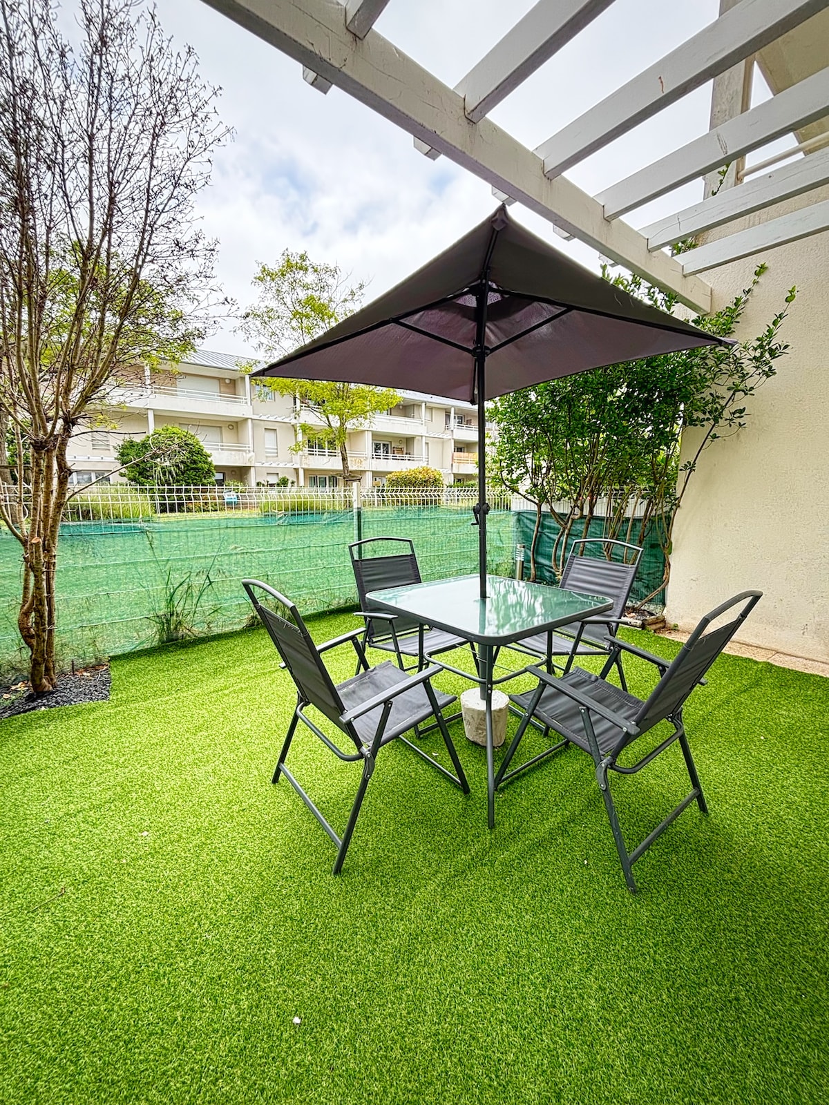 An outdoor terrace features a glass-topped table surrounded by four metal chairs, all situated on artificial grass. A large umbrella provides shade, while greenery is present in the background, contributing to a tranquil environment.