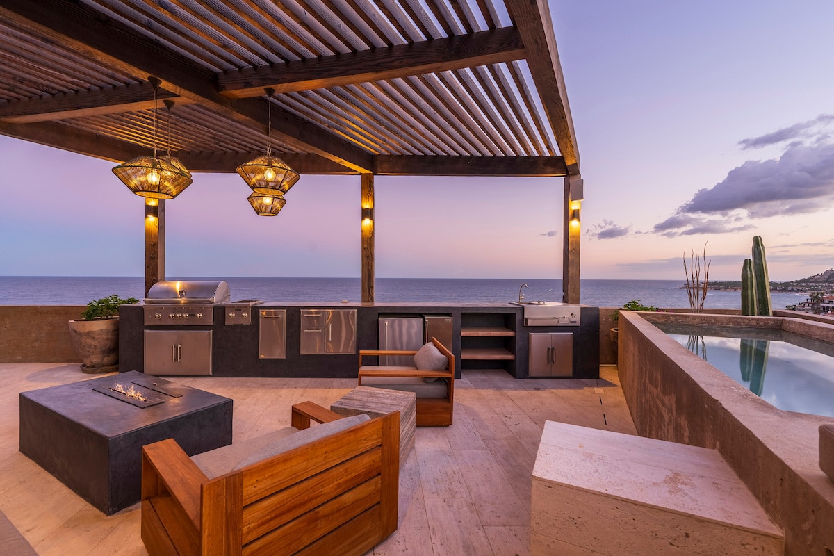 An outdoor kitchen area is showcased, featuring a barbecuing station and a modern sink, accompanied by a dining set. A fire pit sits at the center, while ocean views are visible in the background. Soft light from hanging fixtures illuminates the space as twilight sets in.