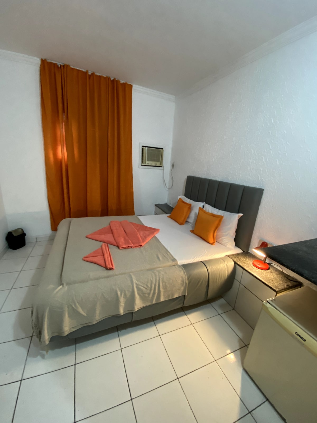 A cozy suite is featured, accommodating two guests. A spacious bed with decorative pillows is set against a light-colored wall. An orange curtain allows natural light, while a small air conditioning unit and a mini-fridge are visible in the room.