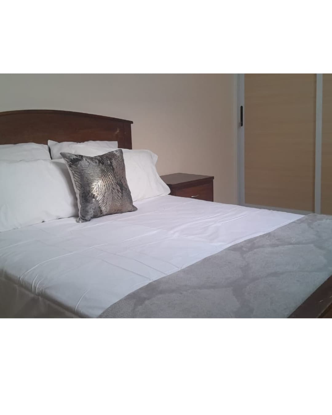 The sleeping area features a spacious bed with crisp white linens and a decorative throw pillow with a subtle sheen. A nightstand is positioned beside the bed, complementing the warm wooden tones of the headboard. Soft natural light filters into the room.