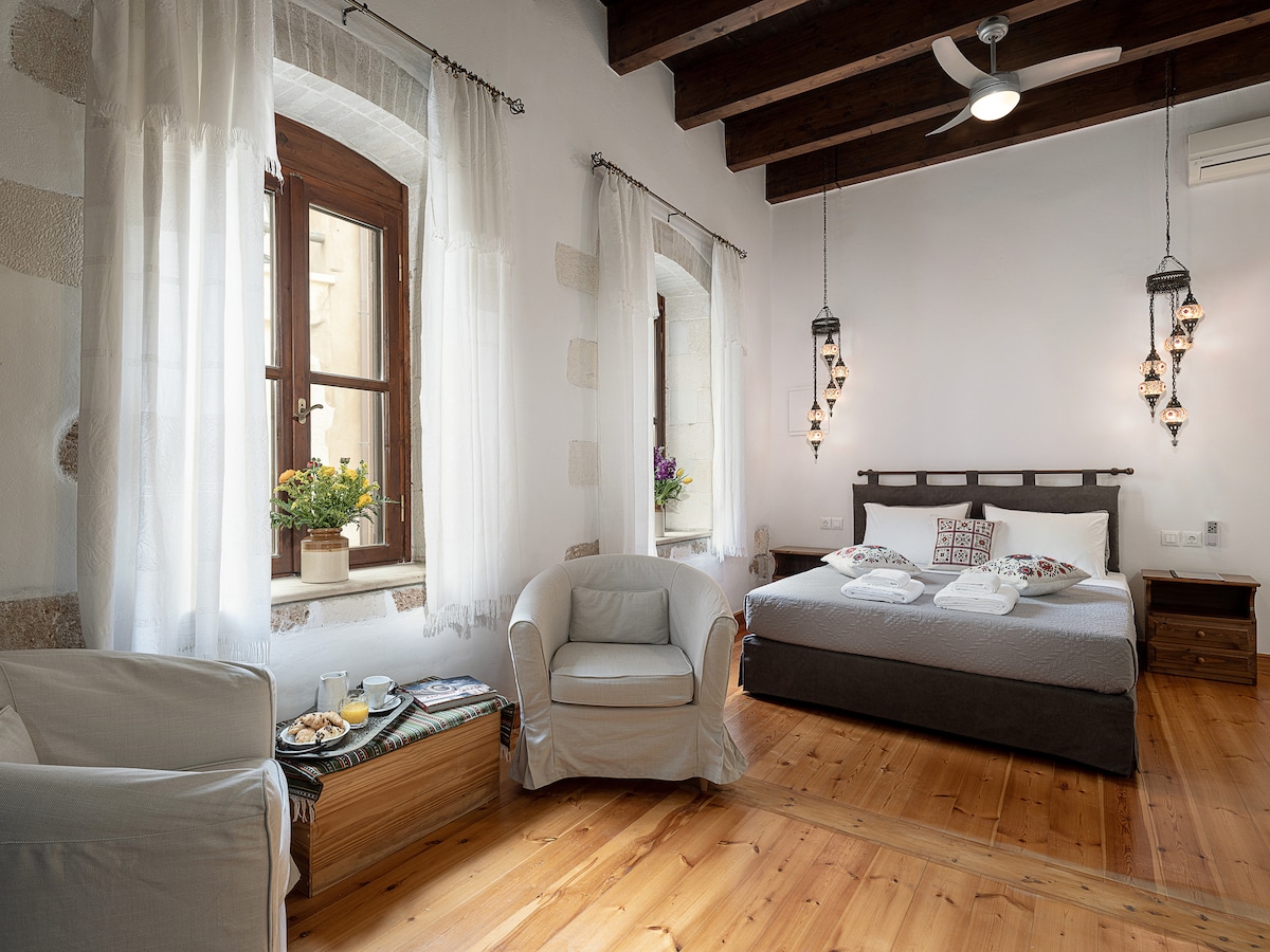 A spacious suite is presented with natural wood accents and light-colored walls. Soft furniture, including a cozy armchair, is arranged by a window draped in sheer curtains. A comfortable bed with neutral bedding sits centrally, complemented by decorative light fixtures hanging from the ceiling.