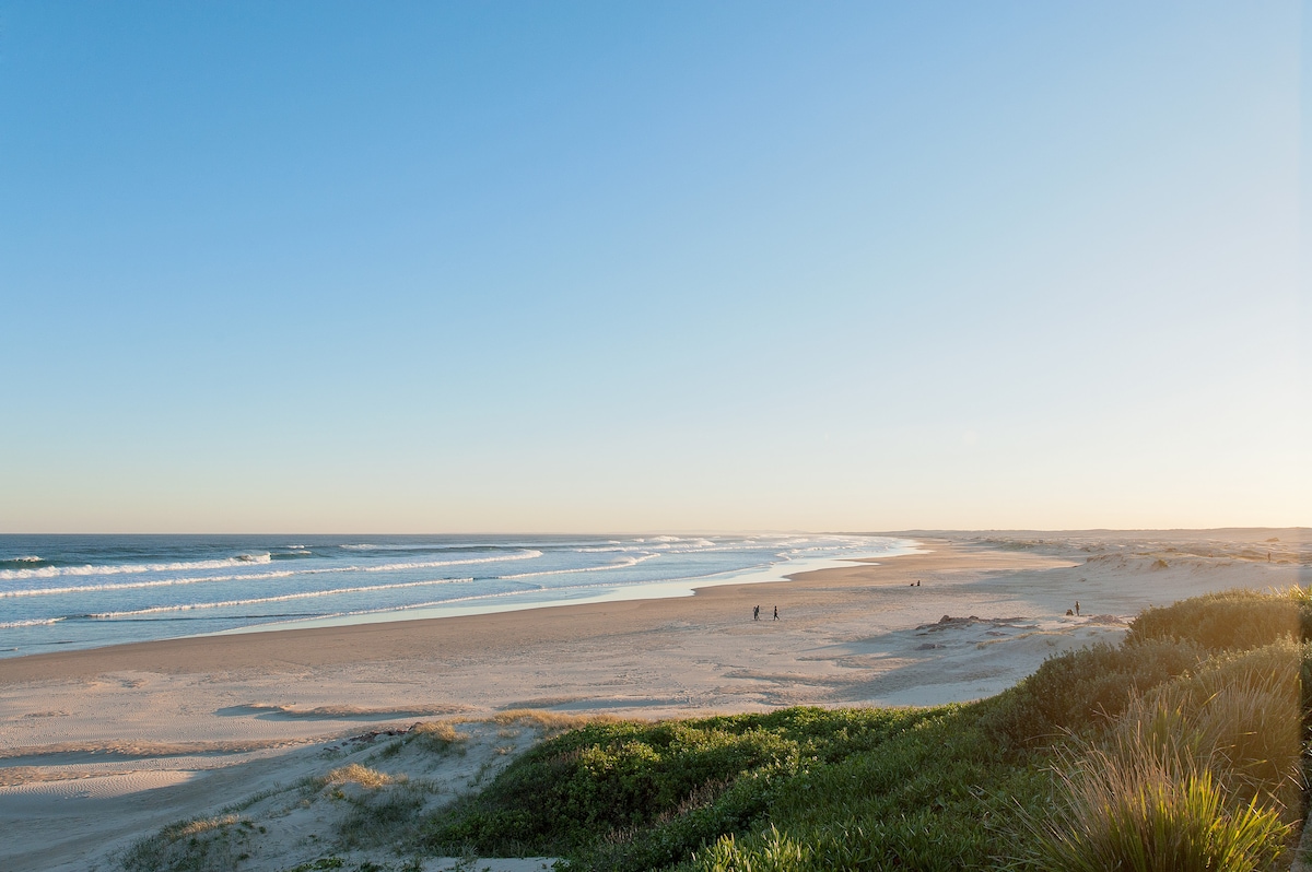 A serene beach scene captures the soft waves lapping at the shore. The expansive sandy coastline is flanked by lush vegetation. People can be seen walking along the water's edge, while a clear blue sky stretches above, suggesting a peaceful atmosphere.
