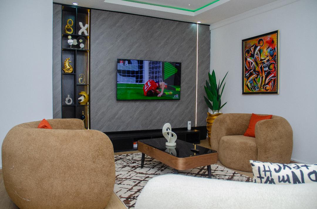 A comfortable living area features a large flat-screen television mounted on the wall. Relaxing round sofas are arranged around a low coffee table, and a decorative bookshelf showcases various art pieces. A vibrant artwork hangs above the seating, adding color to the space.