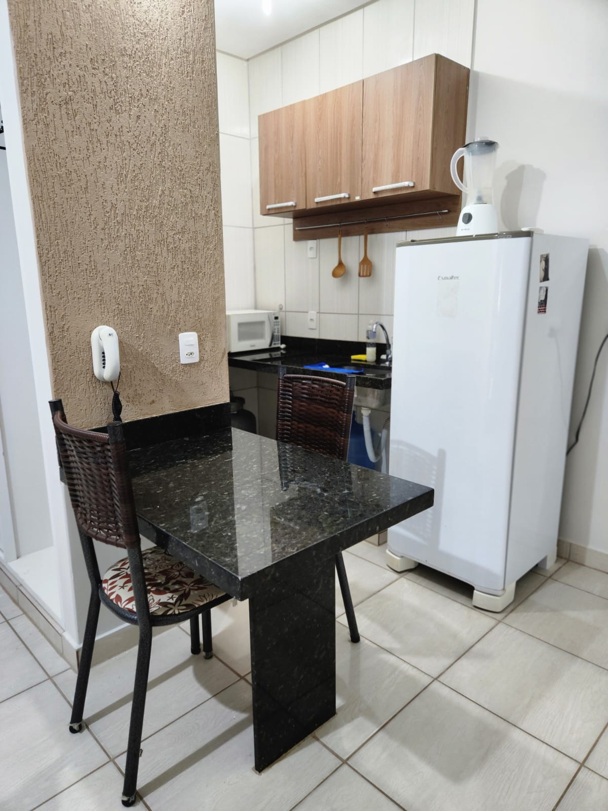 A compact dining area features a dark granite table paired with two woven chairs. In the background, a white refrigerator stands beside a mounted kitchen cupboard, while essential kitchen appliances, including a microwave, are visible on the countertop.