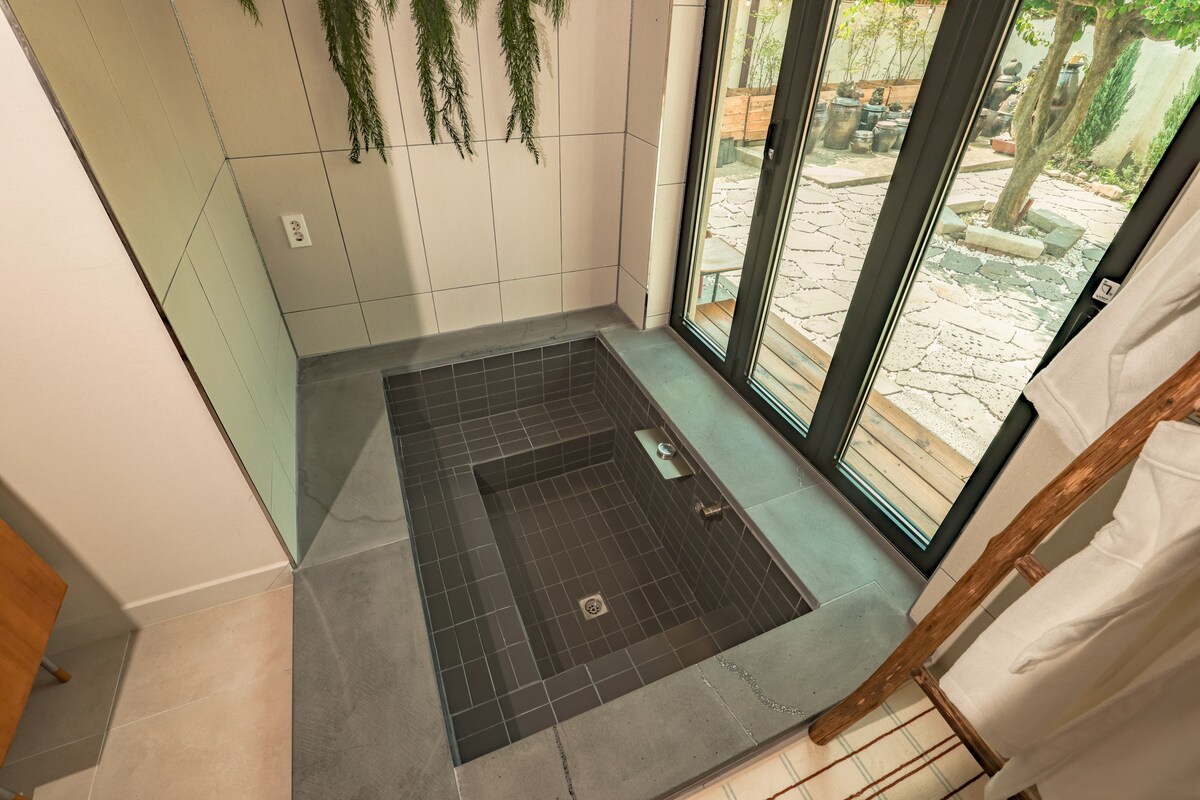 A spacious indoor jacuzzi is set in a sleek, dark tiled area. Natural light filters through large glass doors, revealing a garden view outside. Greenery is displayed on the wall, adding a touch of nature to the relaxing space.