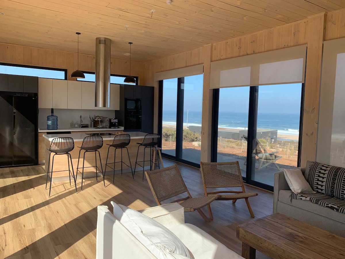 A well-lit living space features a modern kitchen with a central island, complemented by bar seating. Large windows offer expansive views of the ocean. Comfortable seating options include a white sofa and woven chairs, enhancing the sense of openness and connection to the outdoor landscape.
