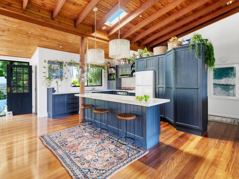 A spacious kitchen features a large island with three woven bar stools, surrounded by blue cabinetry and wooden accents. Natural light floods the area through large windows, highlighting the polished wooden floors and decorative elements. A colorful area rug adds warmth to the design.