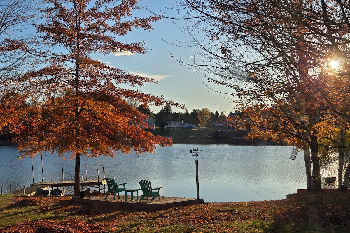 The serene lakefront is framed by trees displaying vibrant autumn foliage. Comfortable seating options in the foreground invite relaxation, while calm water reflects the colors of the trees. A dock is visible, enhancing the setting's connection to the surrounding nature.