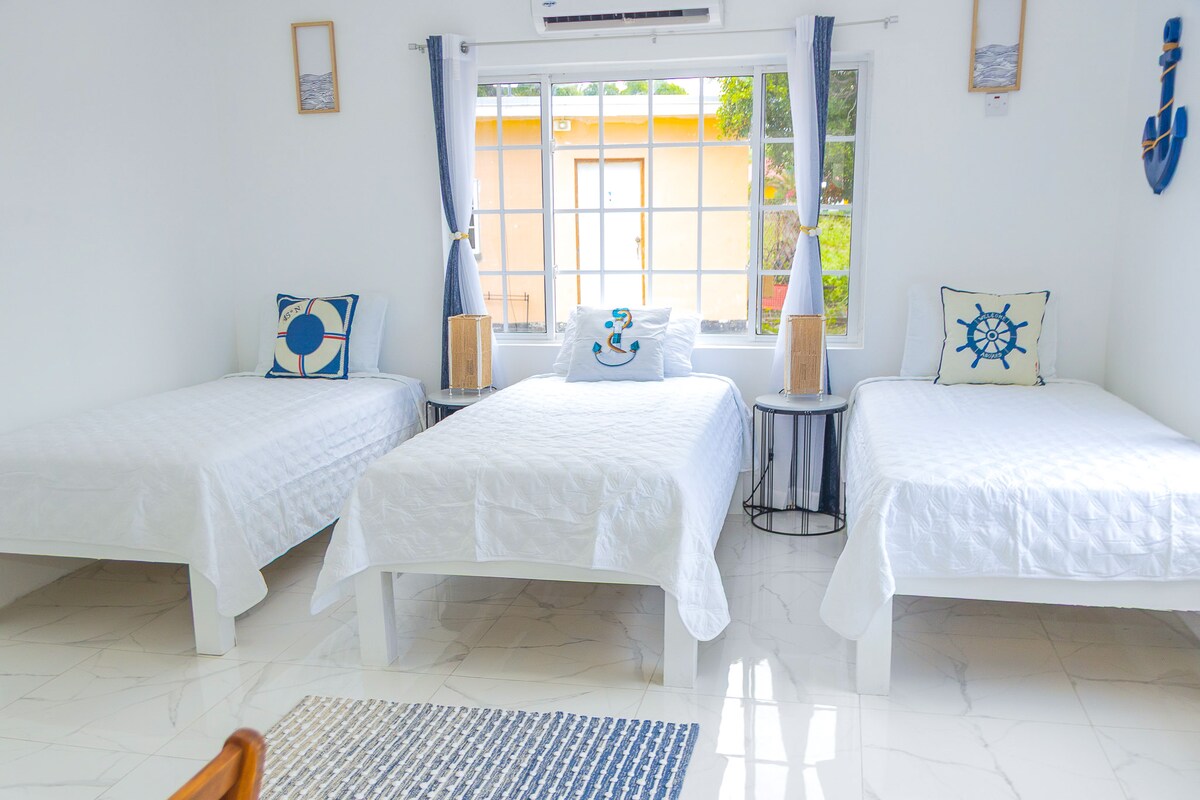 A bright and airy room features three twin beds with white linens arranged neatly. Decorative pillows with nautical themes are placed on each bed. Natural light fills the space through large windows adorned with light curtains, enhancing the fresh atmosphere.