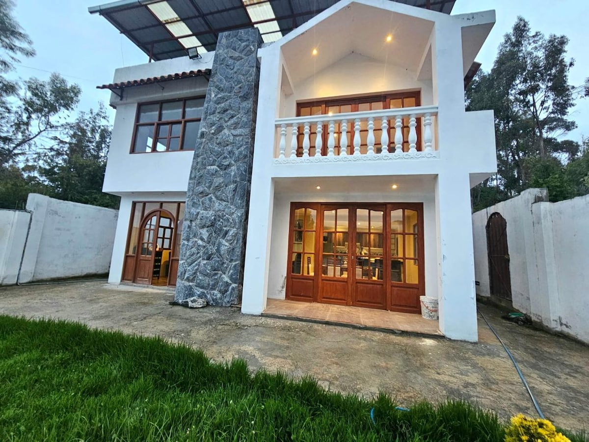 The exterior of a charming two-story house is displayed, featuring a stone accent wall and large wooden windows. An expansive patio is shown, surrounded by well-maintained grass and offering a view of the surrounding natural landscape. A roof provides shelter over the upper balcony.