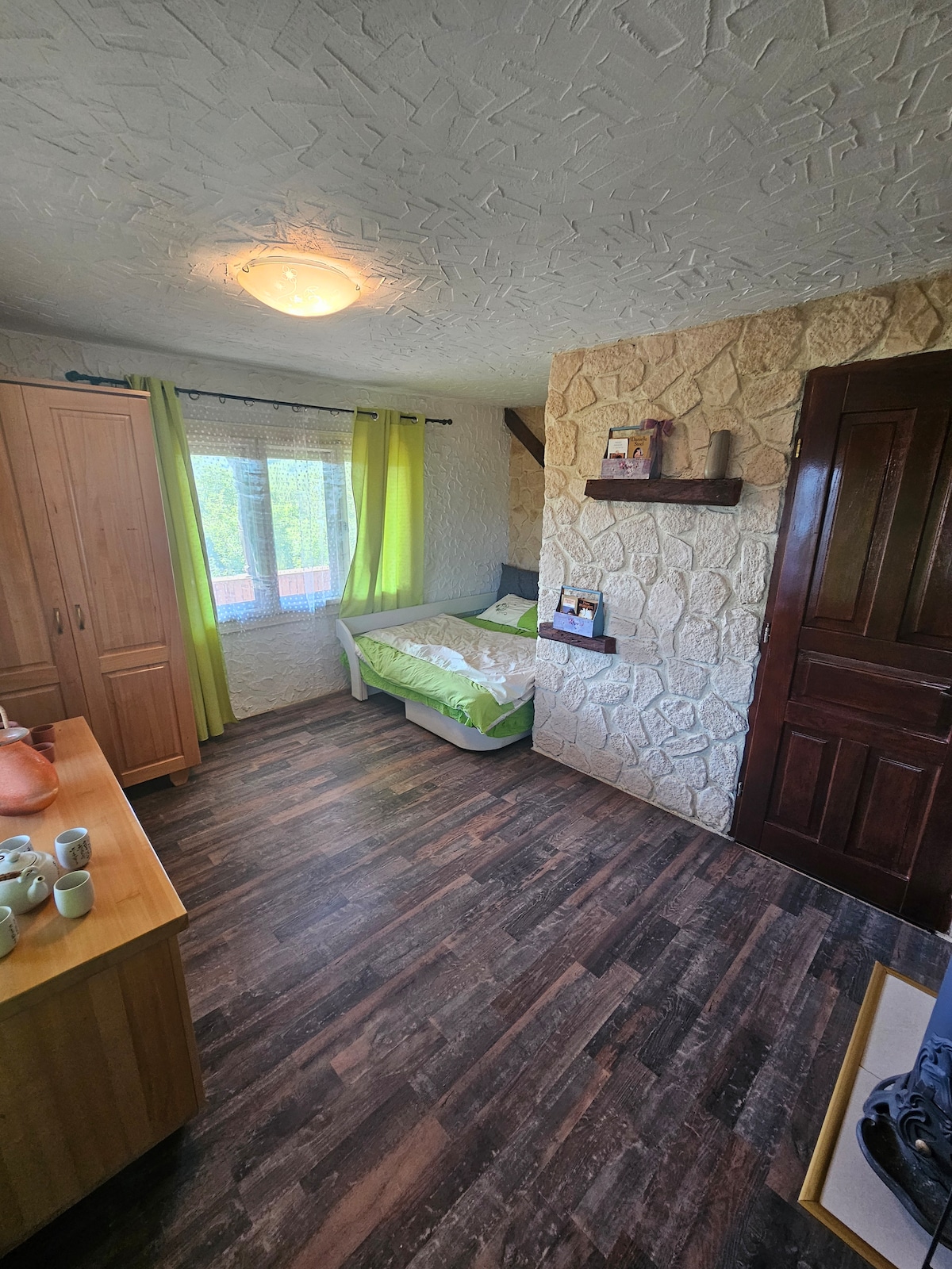 A cozy bedroom features a single bed with a light green duvet, positioned next to a window with sheer curtains allowing natural light to enter. A wooden wardrobe and a dresser with tea cups are visible, along with a textured stone wall and wood laminate flooring.