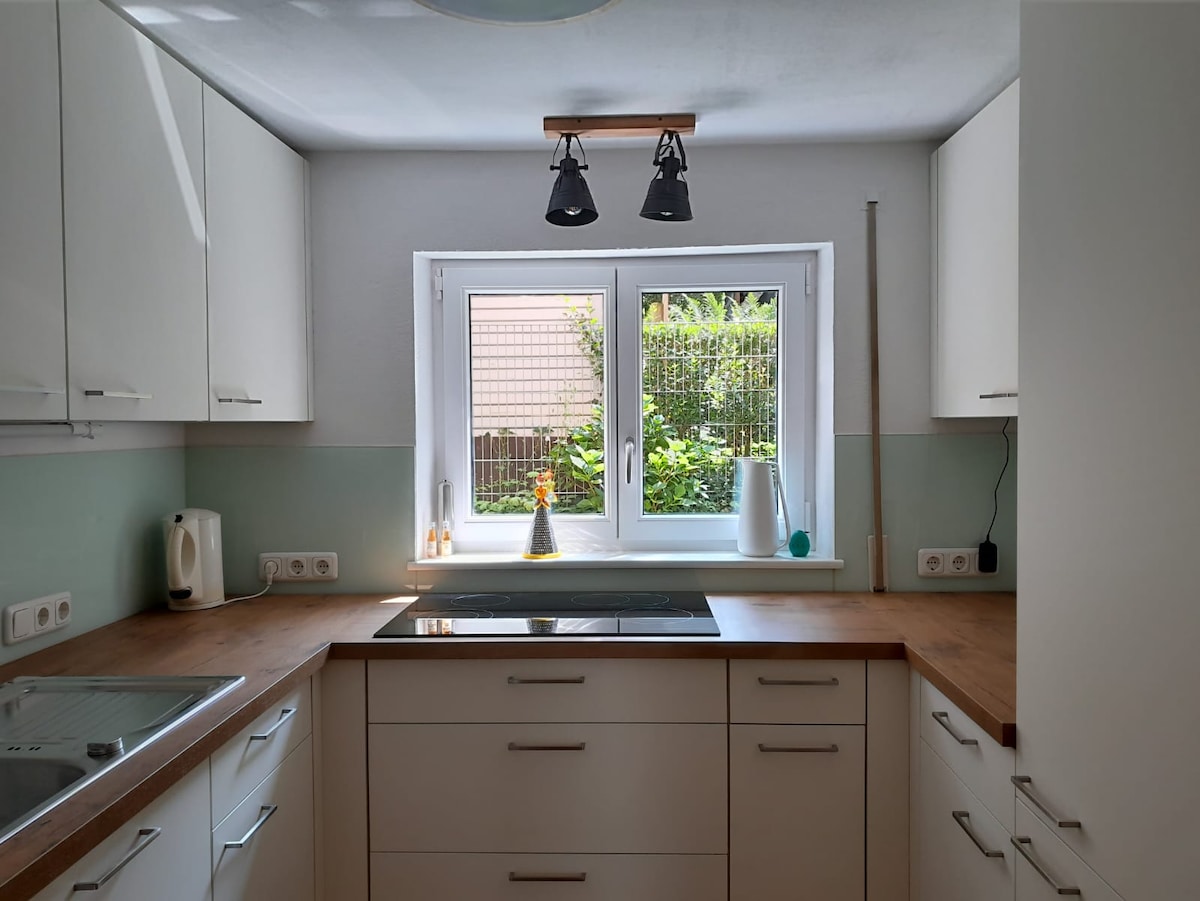 A modern kitchen is presented with light wood countertops and sleek white cabinetry. A window offers a view of the garden, allowing natural light to brighten the space. The kitchen is equipped with essential appliances and features minimalist decor.