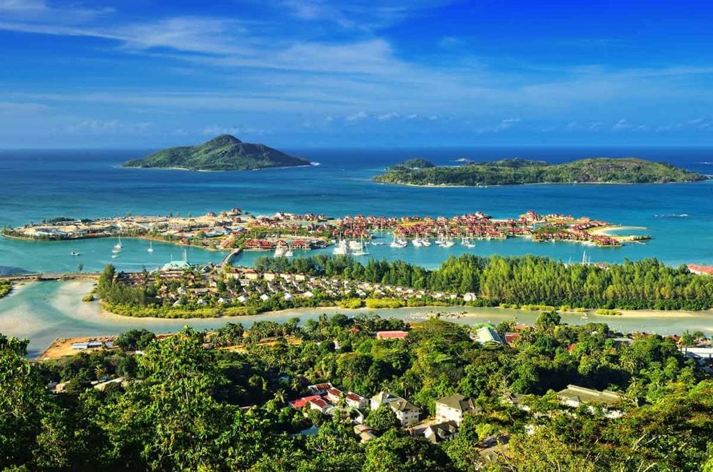 A panoramic view of Eden Island, showcasing a calm bay dotted with boats and surrounded by lush greenery. The tranquil blue waters contrast with the vibrant colors of the buildings along the shore. Islands and hills are visible in the distance, creating a serene coastal landscape.