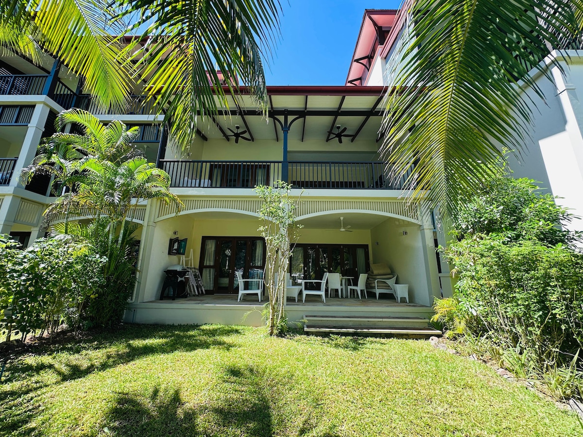 A spacious outdoor area features a grassy lawn surrounded by tropical plants. The building's patio, furnished with a table and chairs, provides shaded seating. Ceiling fans are visible overhead, and a barbecue grill is positioned nearby. Sunlight filters through the greenery.