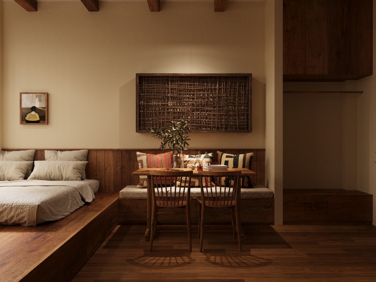 A spacious sleeping area is complemented by a comfortable bed dressed in soft linens. A simple dining table with wooden chairs is positioned nearby, and natural elements are highlighted through a decorative wall piece. Warm lighting enhances the welcoming ambiance of the studio.