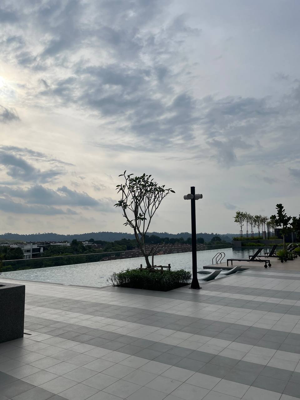 An outdoor area features a serene pool that stretches across the horizon, framed by a stylish infinity edge. A lone tree stands beside the pool, with lounge chairs arranged nearby, creating an ideal space for relaxation amidst a scenic backdrop.