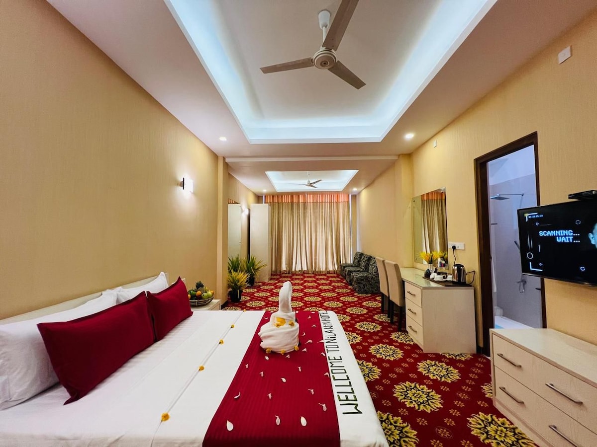 A spacious hotel room is highlighted by a large bed adorned with a deep red and white coverlet. Soft lighting enhances the warm tones of the beige walls. A seating area, coffee station, and television are positioned along one side, while bright curtains frame the windows.