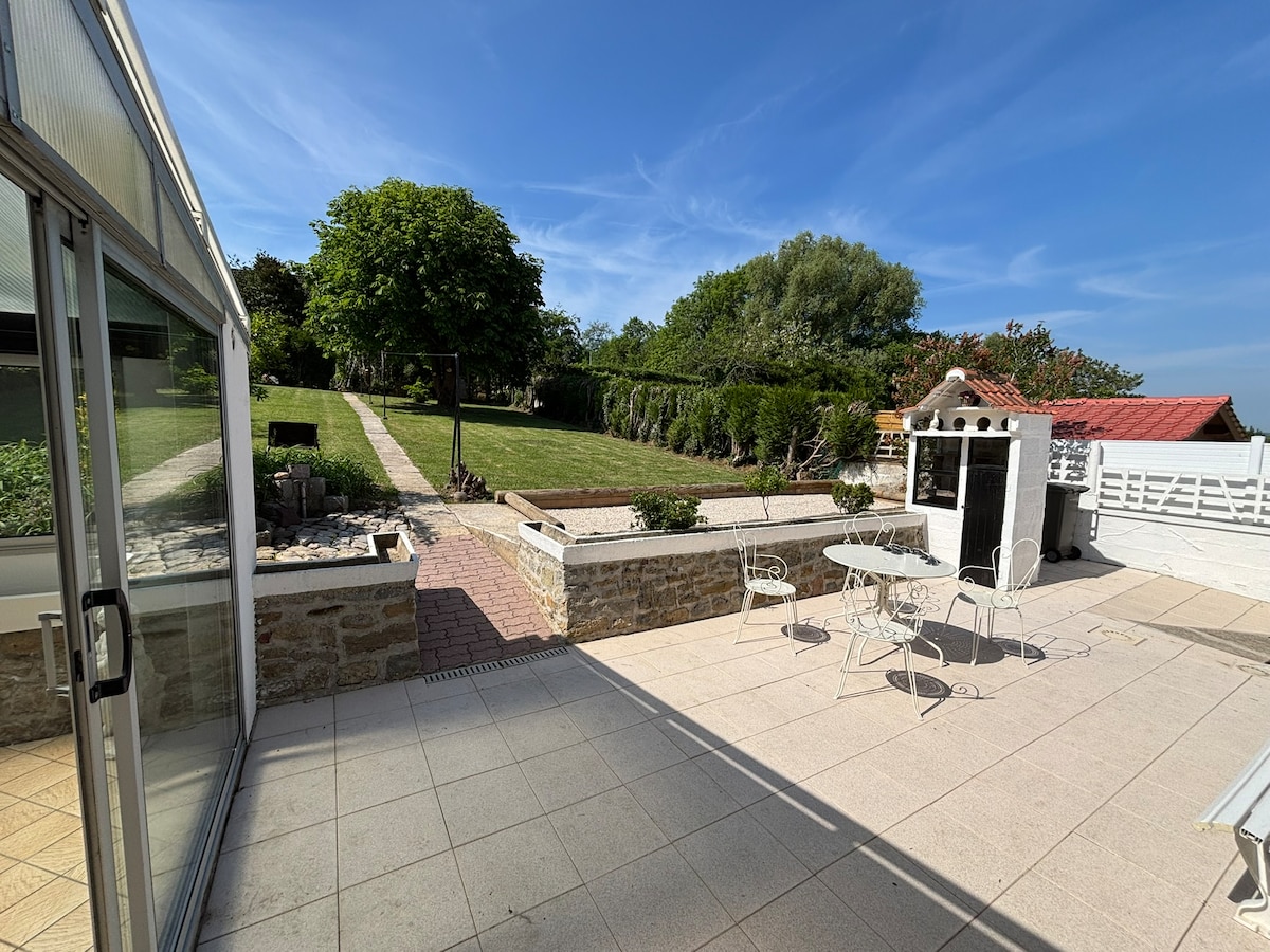 A spacious outdoor area features a paved patio with white metal tables and chairs, set against a backdrop of a lush green garden. A barbecue structure and a stone path lead towards the well-maintained lawn, bordered by trees and garden beds, under a clear blue sky.