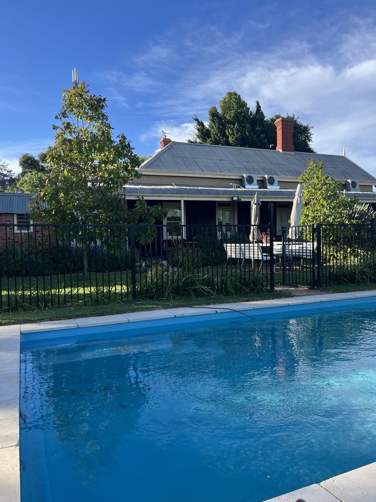 A large swimming pool is visible in the foreground, reflecting the clear blue sky. In the background, a charming old home is surrounded by greenery, featuring a covered porch with umbrellas. Tall trees provide shade, creating a peaceful outdoor setting.