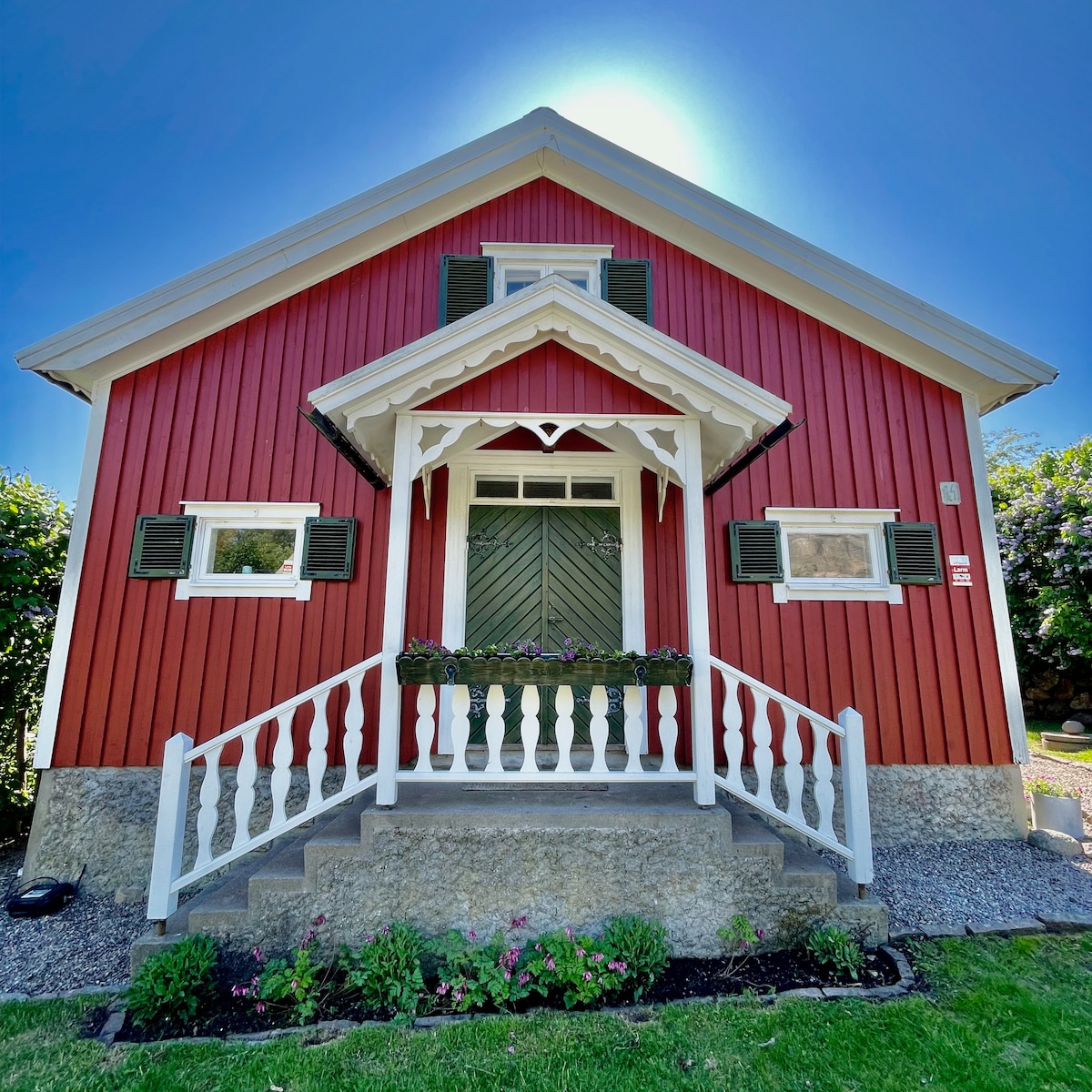 A charming red house features a gabled roof and white trim, with shutters framing the windows. A decorative porch is accentuated by a rail and steps leading to a green door. Lush greenery and colorful flowers are present in the surrounding yard.