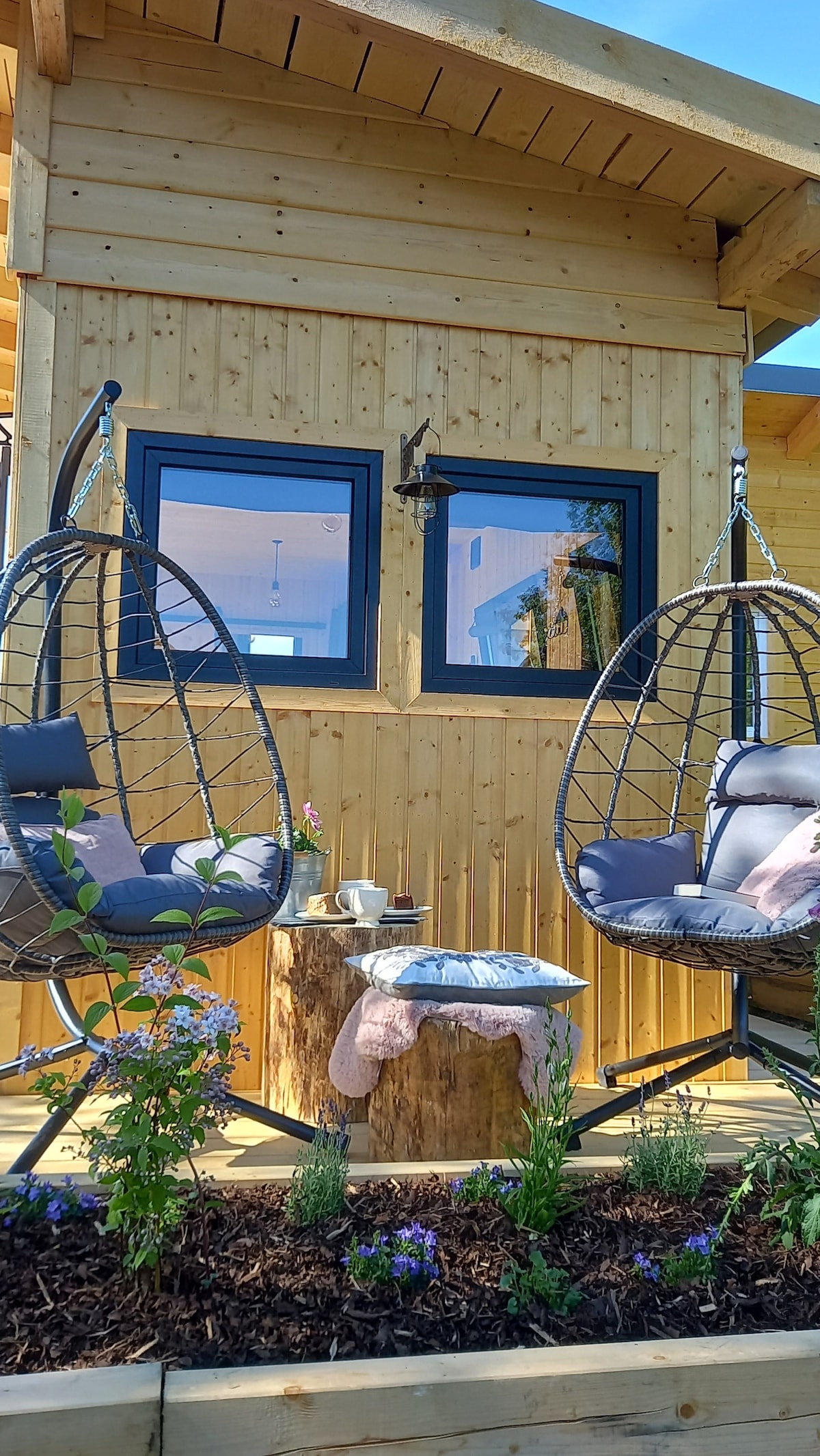 Two hanging chairs are positioned on a wooden terrace, accompanied by a small rustic table. A variety of plants, including flowers, add a natural touch. Large windows on the tiny house allow for natural light, creating a serene outdoor lounging area.