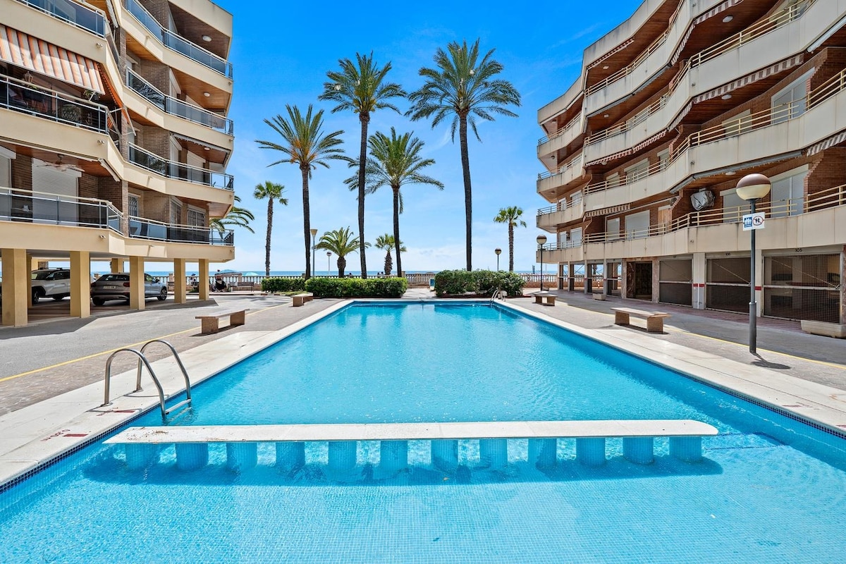A refreshing shared pool is surrounded by palm trees and two residential buildings. The pool area is bright and sunny, with lounge chairs positioned nearby for relaxation. The ocean is visible in the distance, enhancing the peaceful outdoor environment.