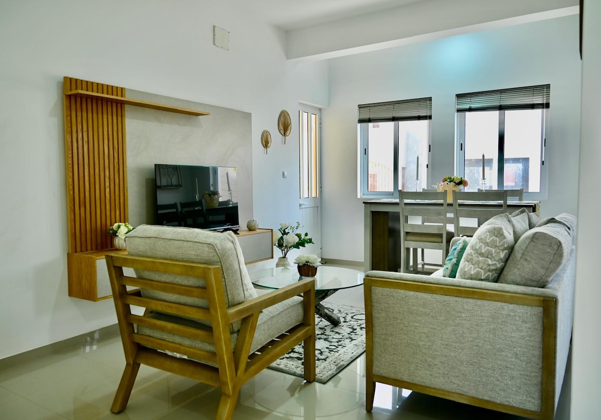 A modern living space features two wooden-framed seating arrangements facing a sleek television mounted on a decorative wall. Natural light enters through two large windows, with a dining area visible in the background, enhancing the open ambiance.