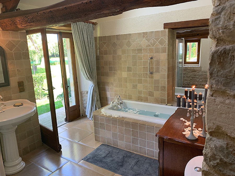 The bathroom features a luxurious spa bathtub surrounded by decorative candles and stone-tiled walls. Natural light enters through the double doors, leading to the garden. A stylish wooden vanity and a round mirror are visible, enhancing the serene atmosphere.