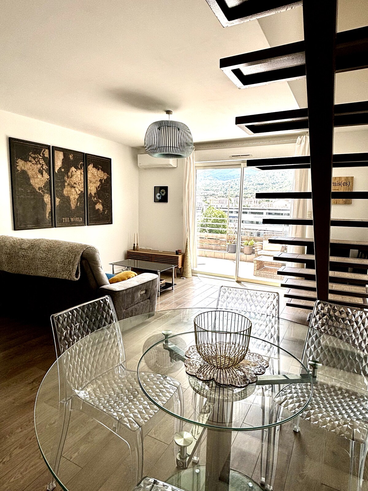 A dining area features a glass table surrounded by transparent chairs. Above, a contemporary light fixture adds an elegant touch. A staircase leads to an upper level, while large windows offer a view of the outside, bringing natural light into the space.