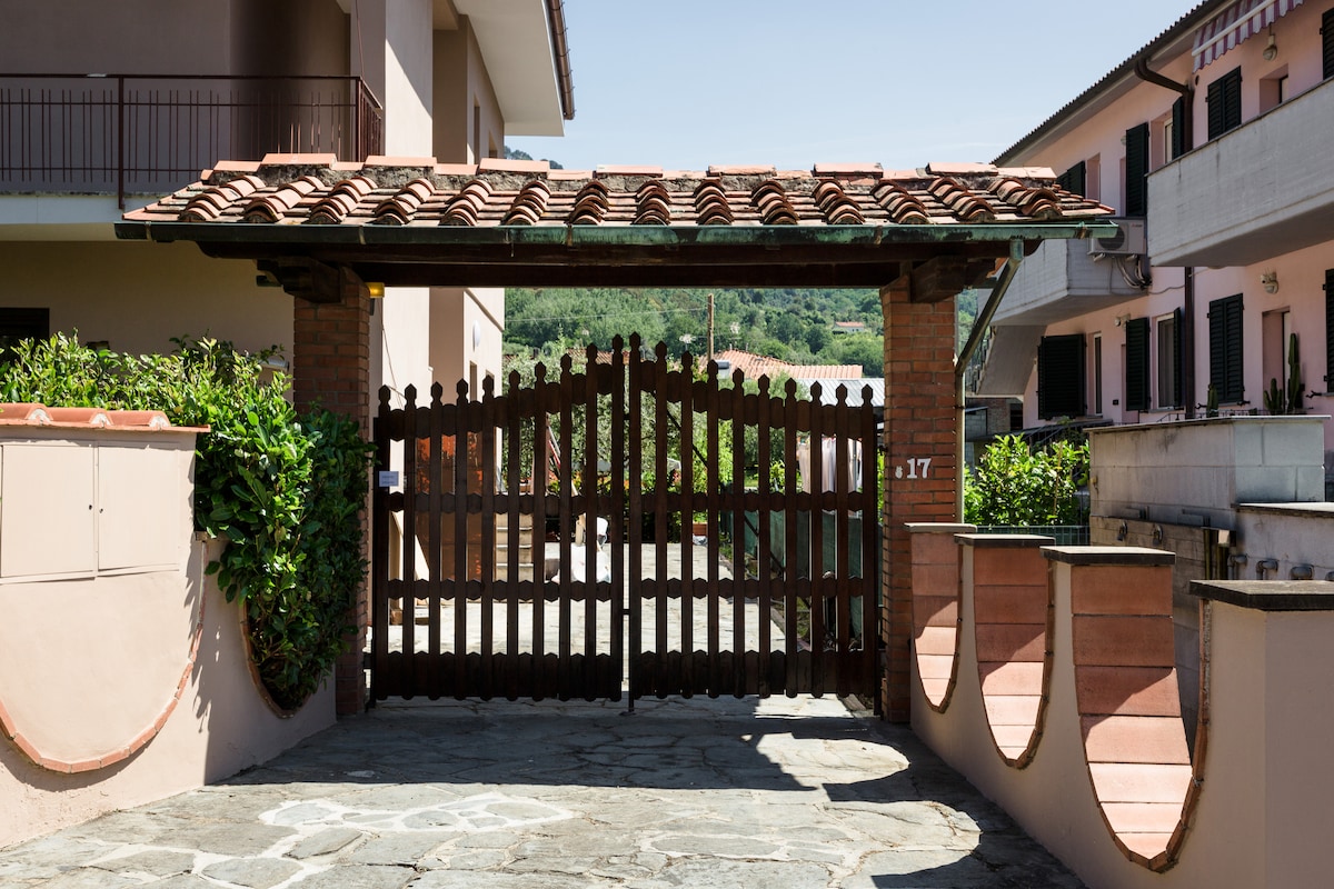 A rustic wooden gate, featuring intricate designs, is framed by terracotta tiles and low stone walls. Lush greenery surrounds the entrance, providing a natural contrast to the earthy tones of the structure. The surrounding buildings are visible in the background, hinting at a welcoming community.
