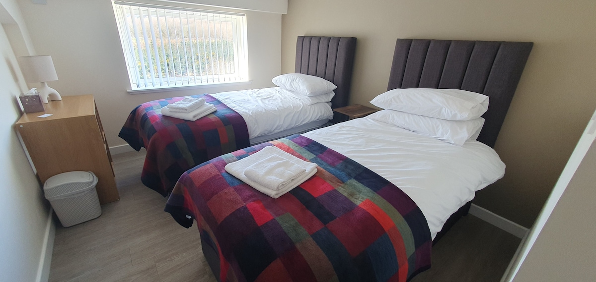 A well-lit bedroom contains two twin beds, dressed in white linens and colorful plaid throws. A bedside table is positioned beside each bed, with a small lamp on one. A window with vertical blinds allows natural light to fill the room.