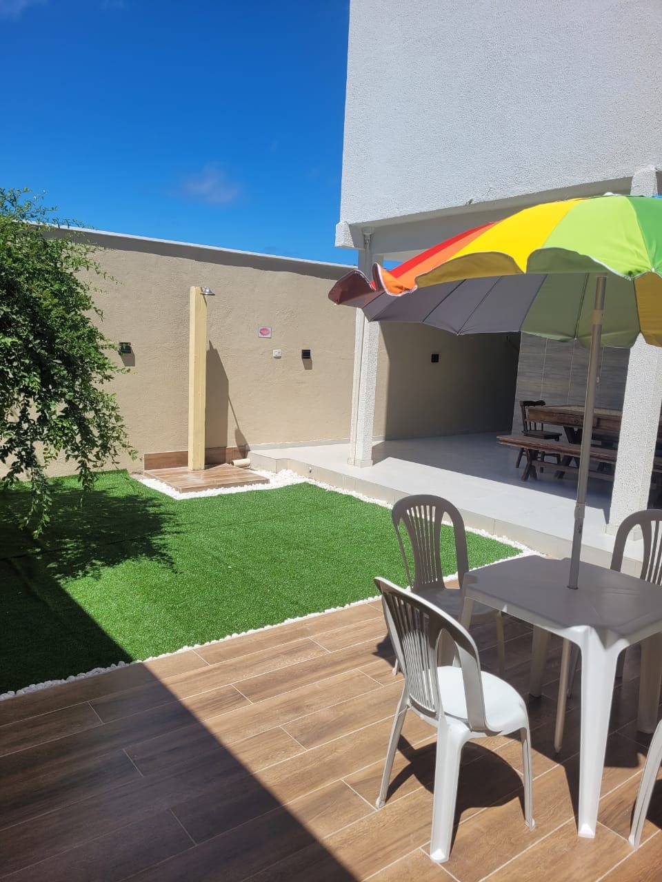 A well-maintained outdoor area features a green lawn and a small shower space surrounded by beige walls. A table with four chairs is shaded by a large, colorful umbrella, while a dining set is visible in the background. Bright blue skies enhance the open atmosphere.