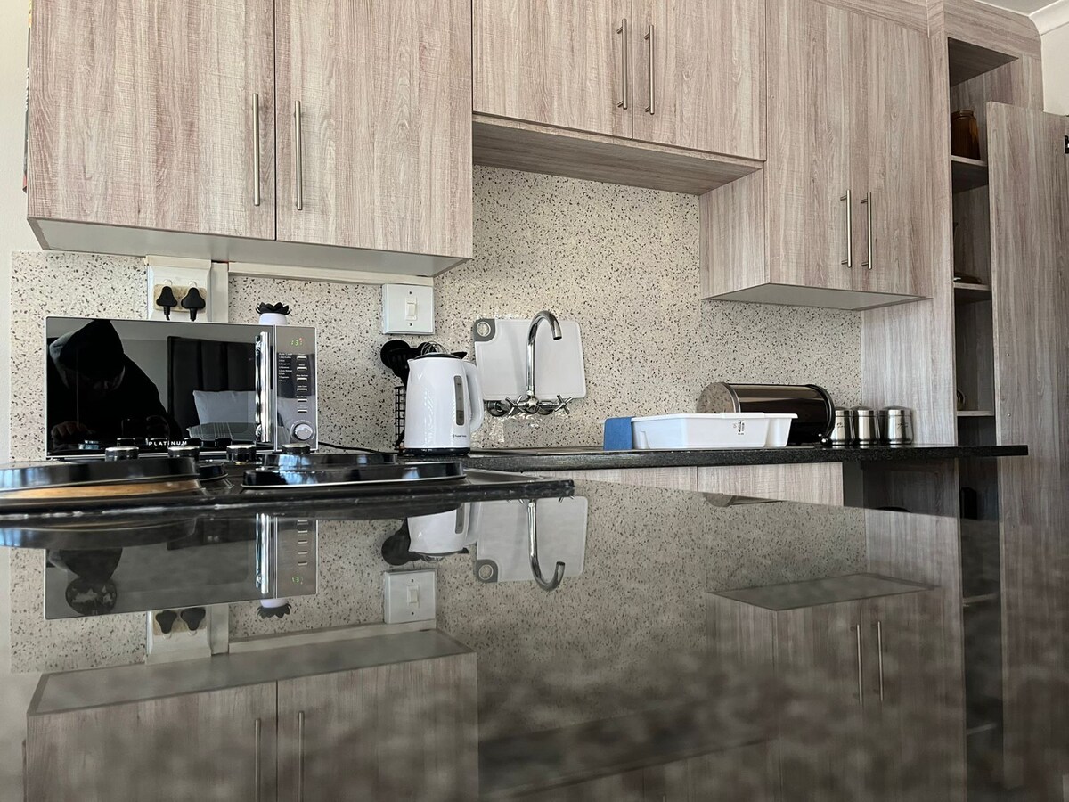 A modern kitchen is highlighted by sleek cabinetry and a polished countertop. Essential appliances, including a microwave and kettle, are positioned conveniently. Natural light reflects off the smooth surface, enhancing the overall clean and organized appearance of the space.