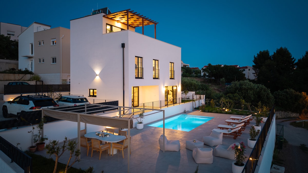 A modern villa is showcased with clean lines and a flat roof. An outdoor pool glistens under the evening sky, surrounded by lounge chairs and a dining area. Cars are parked nearby, and lush greenery adds a natural touch to the landscape.