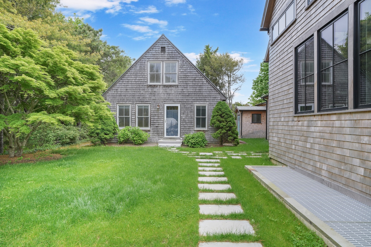 A charming guest house is surrounded by thriving greenery and well-maintained grass. A stone pathway leads to the entrance, with large windows on the front exposing a glimpse of the interior. Mature trees provide natural shade and enhance the serene atmosphere of the property.