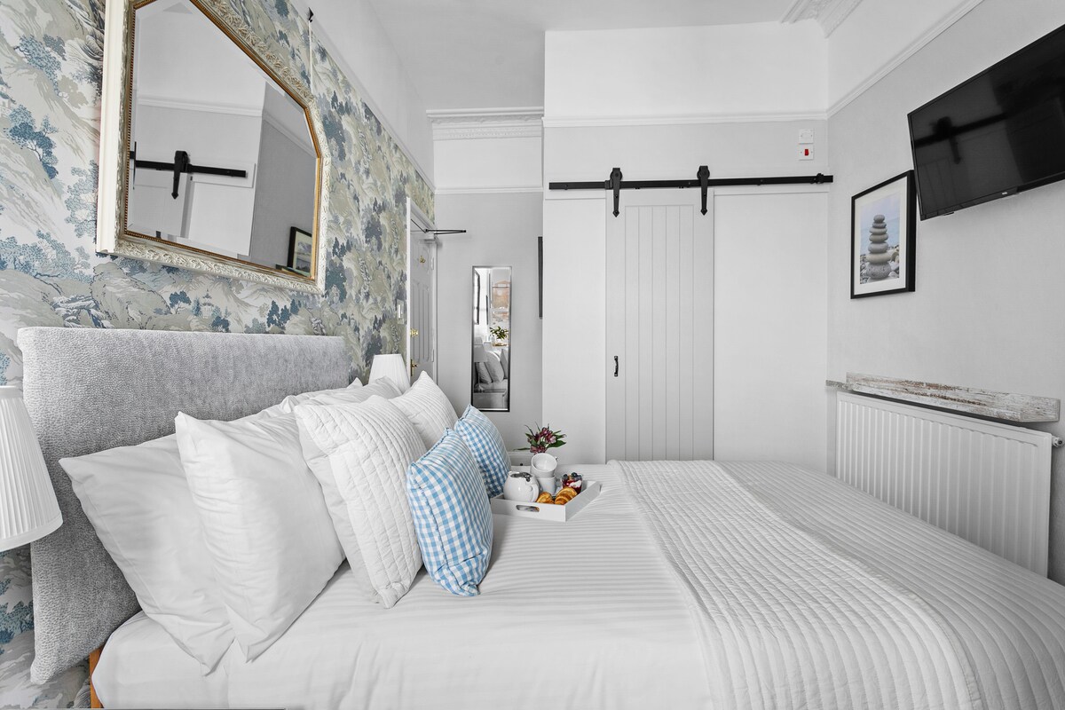 A light and airy room features a spacious bed adorned with crisp white linens and decorative pillows. A small tray with fruits sits at the foot of the bed. The wall is decorated with floral wallpaper, and a mirror reflects the room's stylish decor. A Smart TV is mounted on the wall.